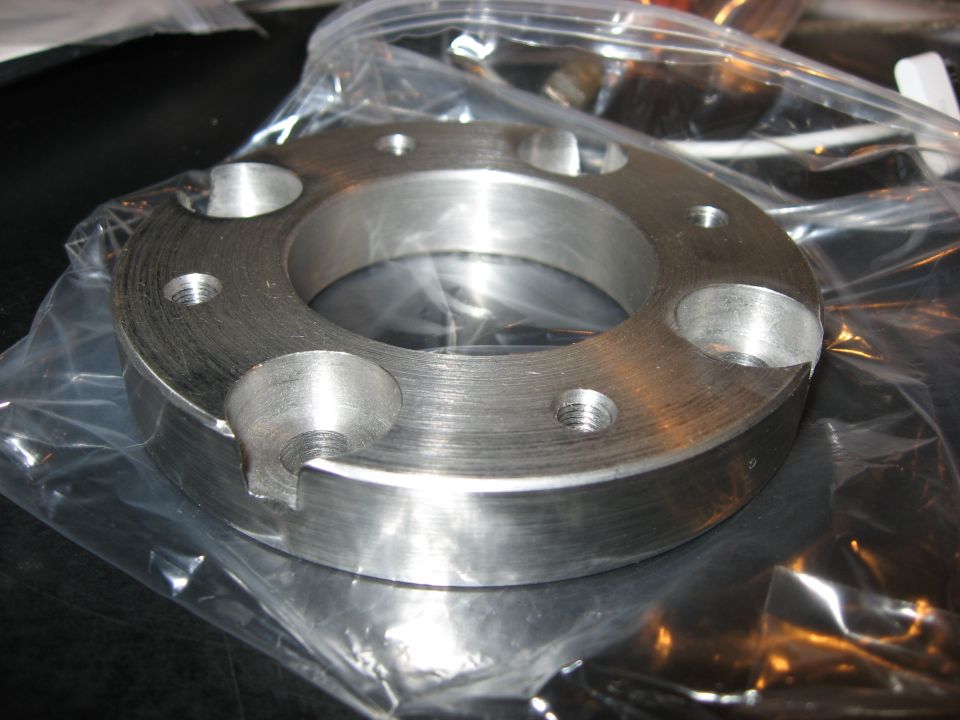 Machined metal spacer with six bolt holes and a central opening, partially in plastic packaging