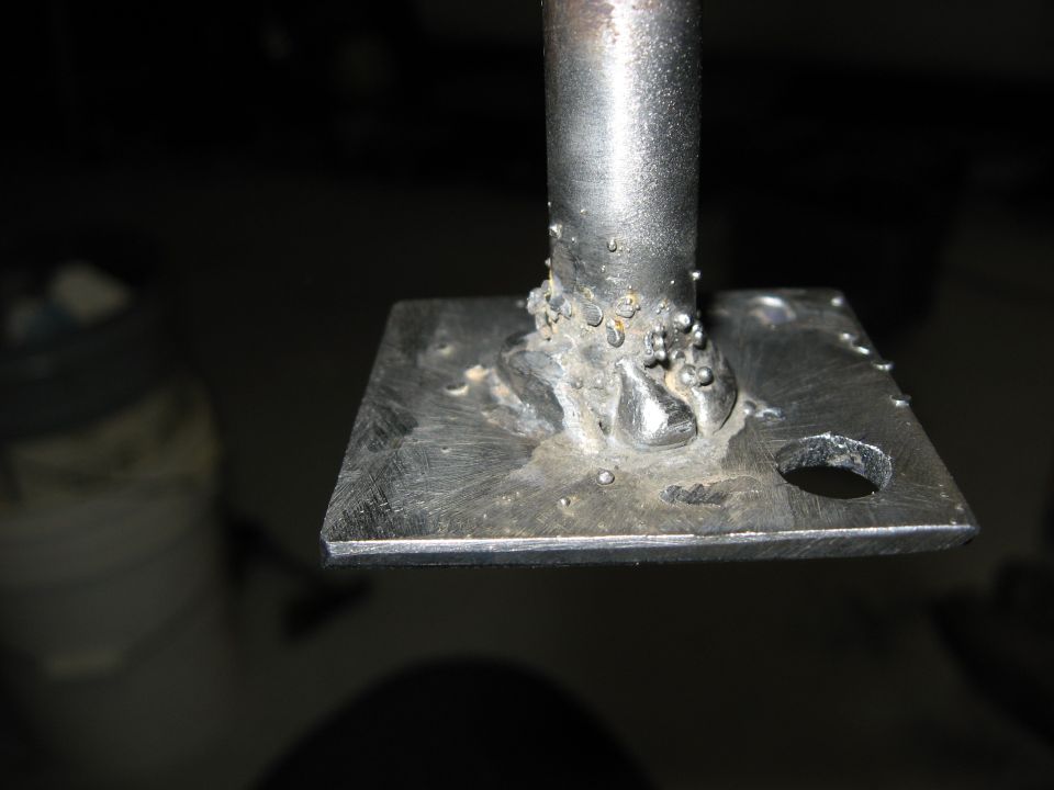 Close-up of a rough metal weld on a square plate with a hole.