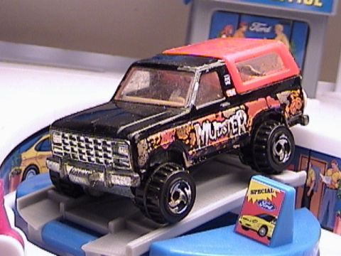 1979 Ford Bronco - A black toy truck with a red top and 'MUDSTER' graphics sits on a display ramp.