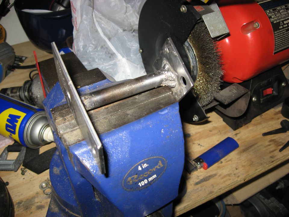 Metal bracket clamped in a blue vise, wire wheel grinder and WD-40 can in background.