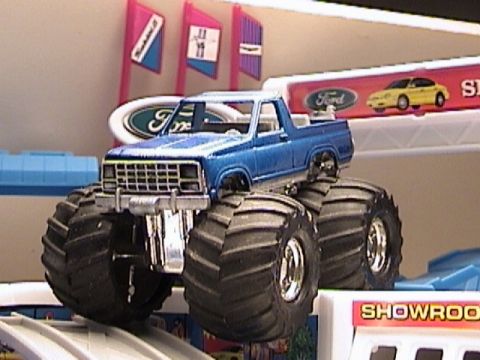 1979 Ford Bronco - This blue 1979 Ford Bronco monster truck is displayed with showroom signage in the background.
