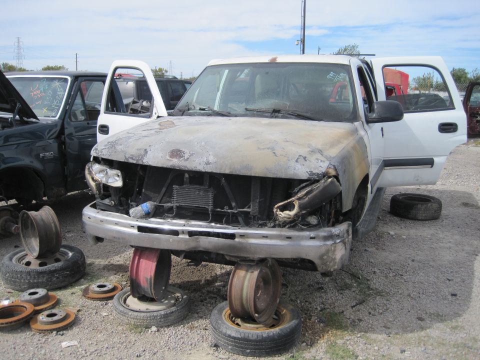 Damaged white pickup truck in junkyard with open hood and destroyed front end.