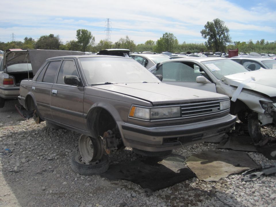 Brown sedan in a junkyard, missing front wheels.