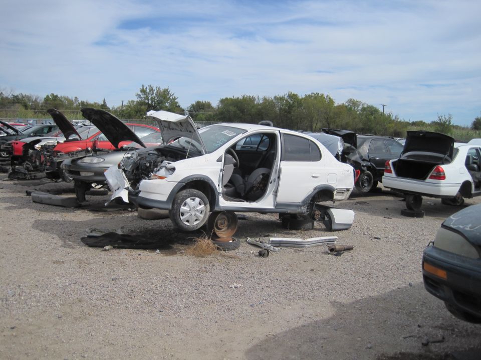 White sedan with hood and doors open, elevated in a junkyard.