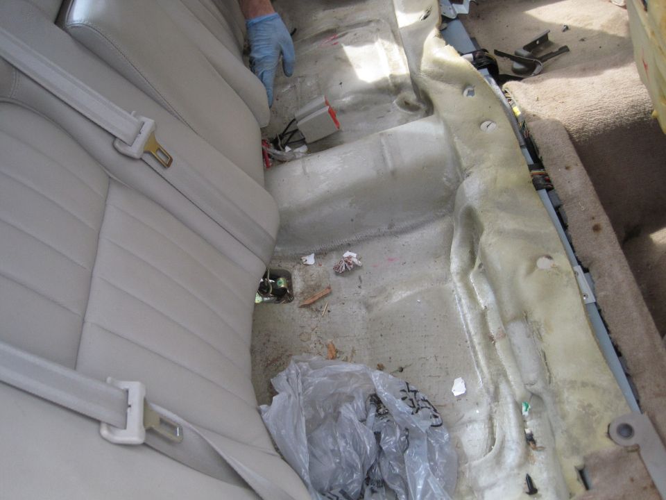 Car interior with rear seat removed, showing floor, wiring, and a gloved hand near seatbelt.