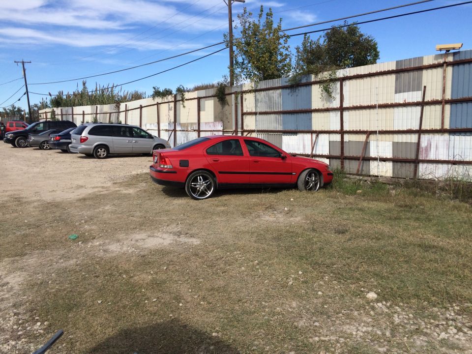 Red sedan with chrome rims parked in front of a corrugated metal fence.
