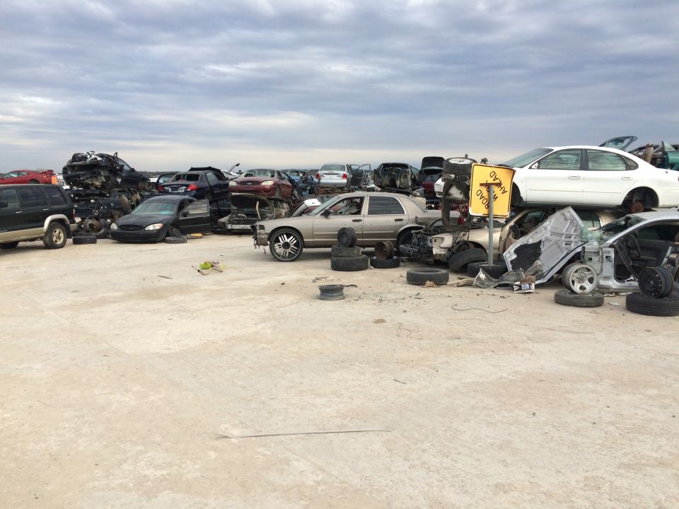 Junkyard with stacked and wrecked cars, foreground silver sedan, cloudy sky.