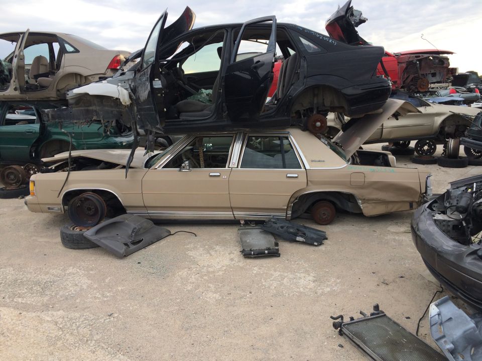 Junkyard scene with a beige sedan in the foreground, other cars stacked on top.