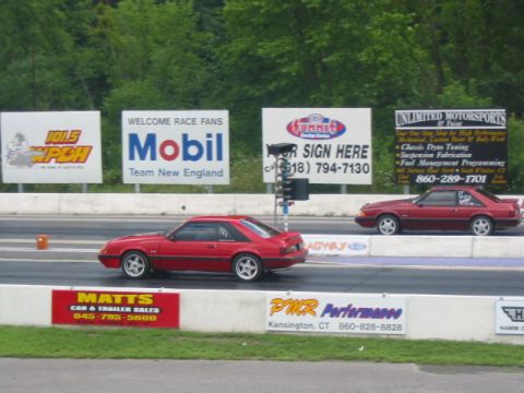 1986 Ford Mustang - Two red 1986 Ford Mustangs compete on a drag strip, with various sponsor advertisements visible.