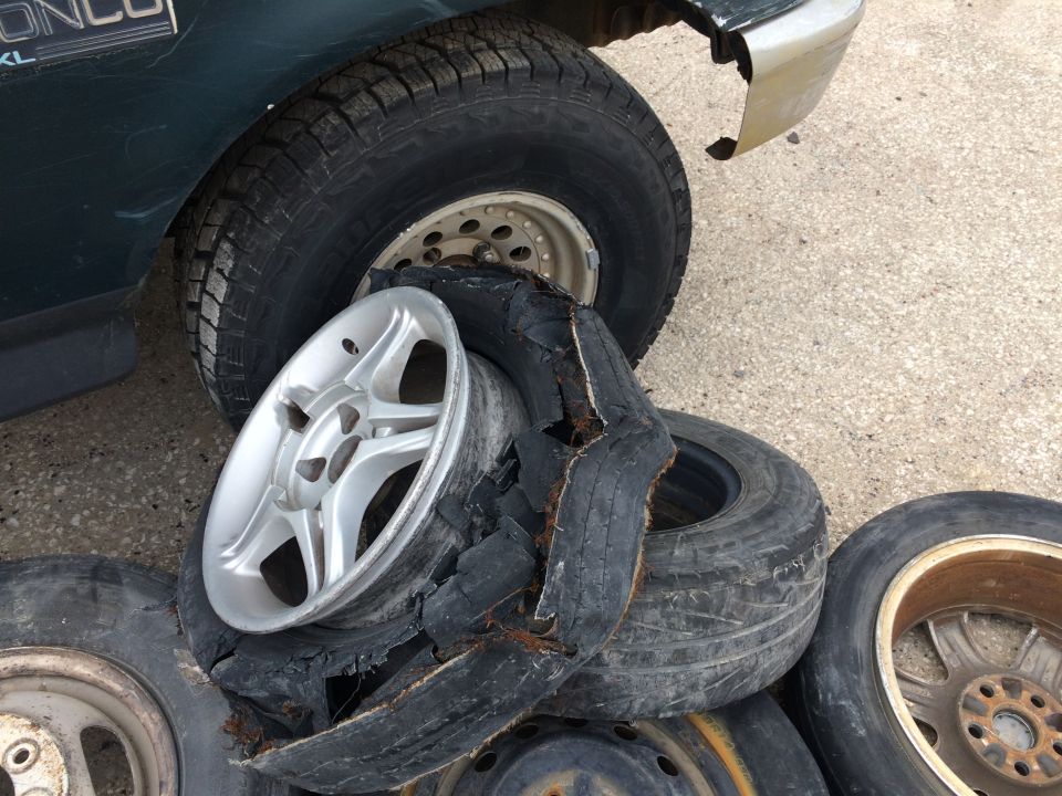 Damaged tire and rim next to a dark green vehicle with other tires and rims piled nearby.