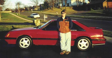 1986 Ford Mustang - A person poses with a red 1986 Ford Mustang coupe parked on a residential street.