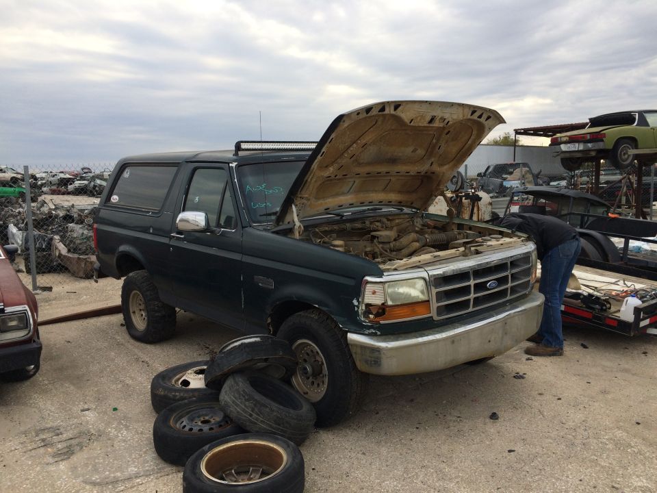 Dark green Ford Bronco with hood open in a junkyard, person looking at engine, tires stacked nearby.