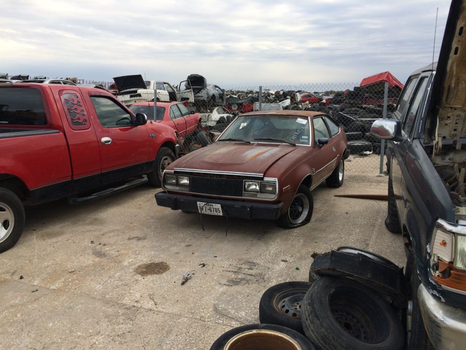 Rusty brown AMC Spirit in a junkyard with a red pickup truck.