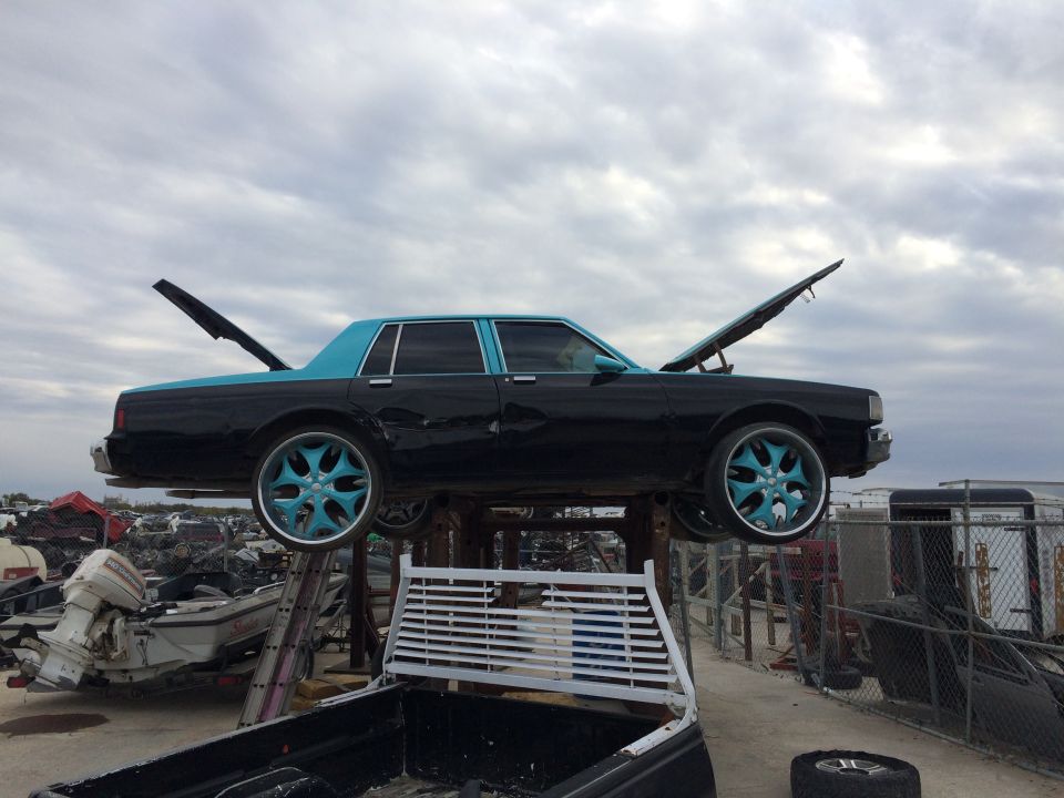 Black and teal sedan with open hood and trunk, custom teal rims, elevated in a junkyard.