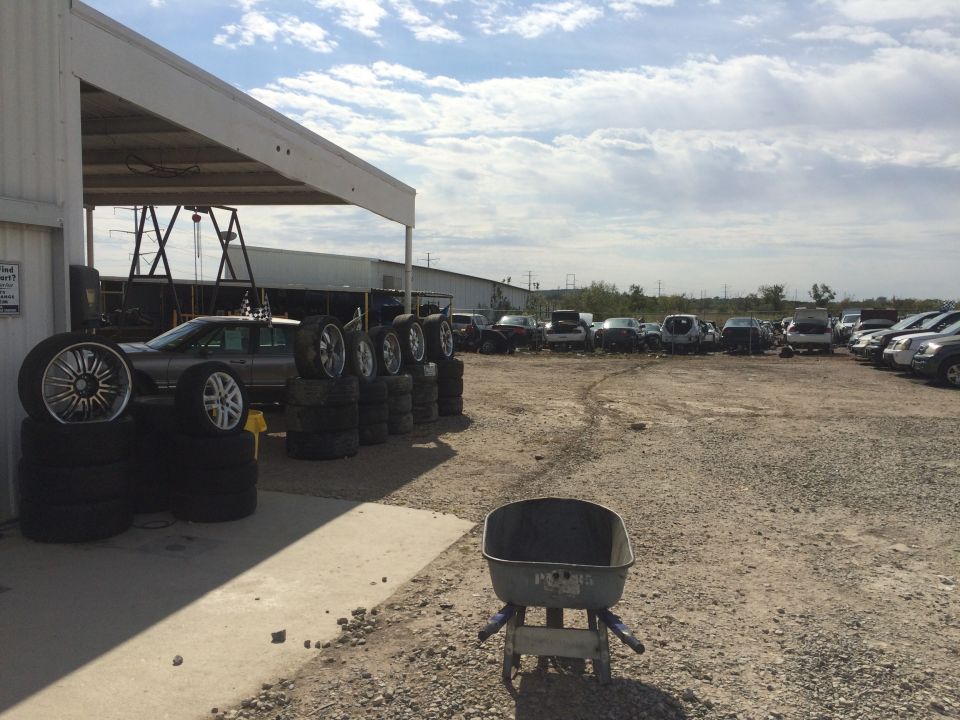 Dark sedan with stacks of tires and rims in front, junkyard in background.