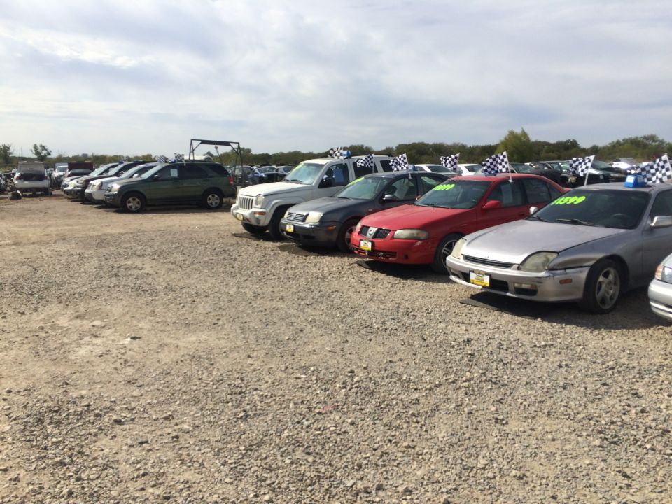 Used cars for sale on a gravel lot, some with checkered flags.