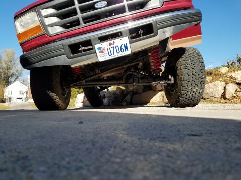 1995 Ford Bronco - Low-angle shot of the front right tire and suspension of a red Ford Bronco. The vehicle is parked on a concrete driveway with a house and t…