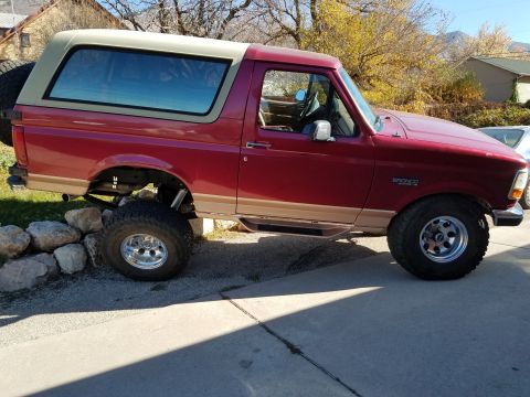 1995 Ford Bronco - A red 1995 Ford Bronco is parked on a concrete driveway with a beige top and tan lower body trim. The vehicle has large off-road tires and…