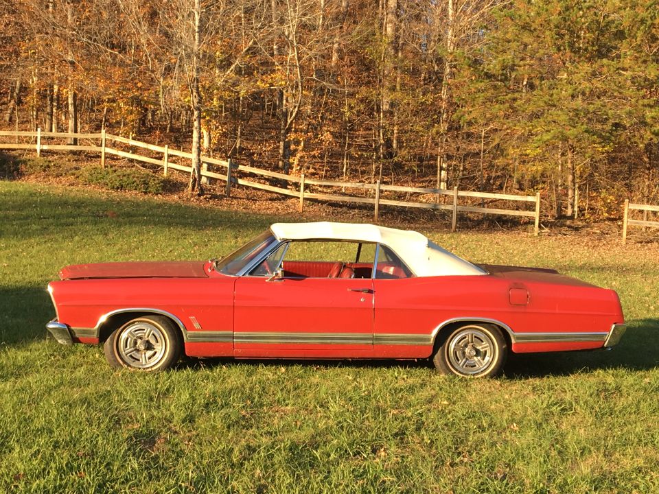 Red 1967 Ford Galaxie convertible with white top parked on grass, autumn trees in background.