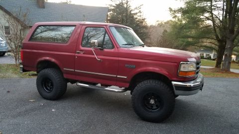 1994 Ford Bronco - A red Ford Bronco is parked on an asphalt driveway in front of a house and trees. The vehicle is shown in a side profile with its front pas…