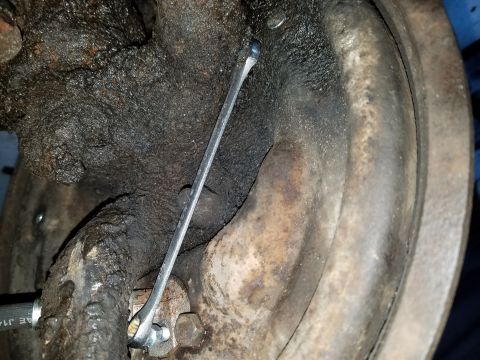 1971 Ford Mustang - A close-up view of a rusty and greasy brake drum assembly, with a wrench positioned between the brake shoes. A portion of a brake hose with…