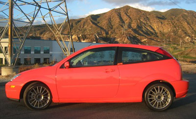 1998 Ford Crown Victoria - Side profile of a red three-door Ford hatchback parked near a low industrial building with mountains in the background.