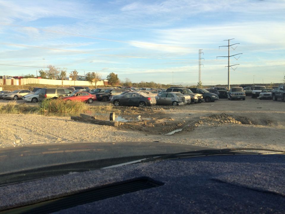 Junkyard scene with numerous vehicles parked on dirt lot under a cloudy sky