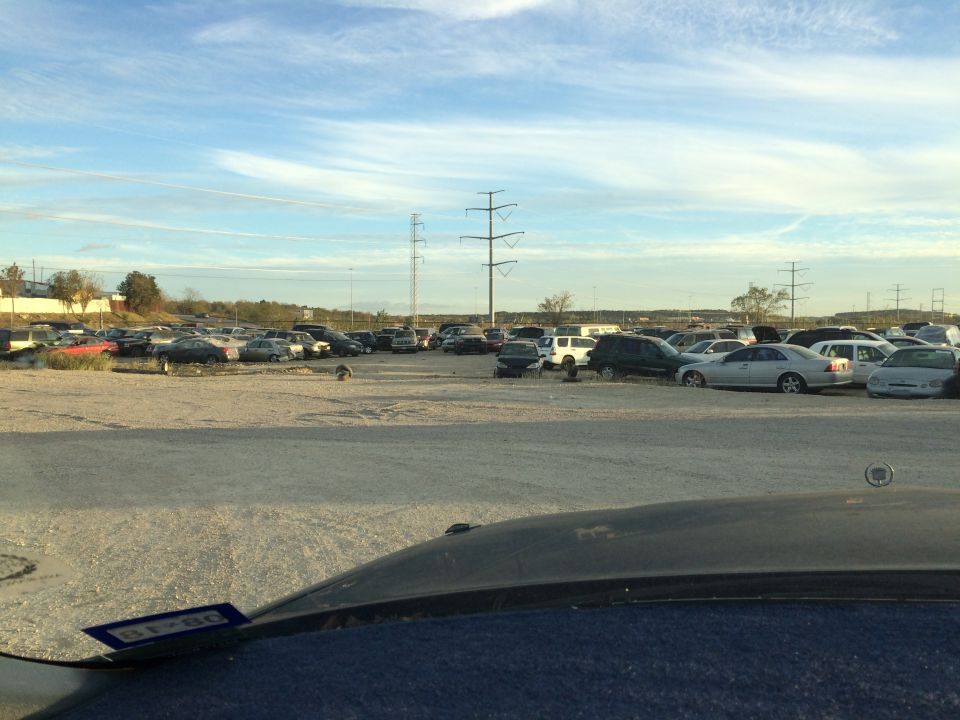 Junkyard scene with many cars under a cloudy sky. A license plate is visible on the dashboard