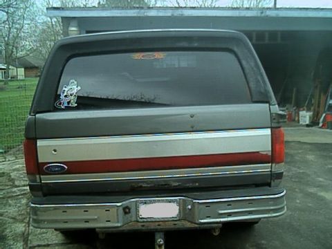 1989 Ford Bronco - Straight-on rear view of a gray full-size Bronco-style SUV with the hardtop in place, showing the tinted rear glass with decals, a red and…