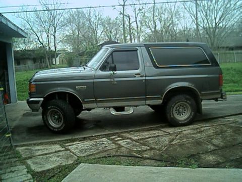 1989 Ford Bronco - Driver-side profile of a two-tone gray full-size Ford Bronco with its hardtop in place, parked on a concrete driveway beside a bare-tree ya…