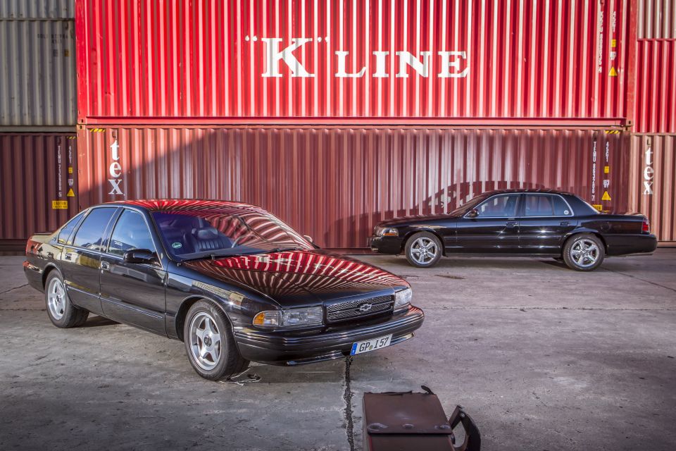 Two black sedans parked outdoors in front of red K-Line shipping containers. One car is closer to the camera