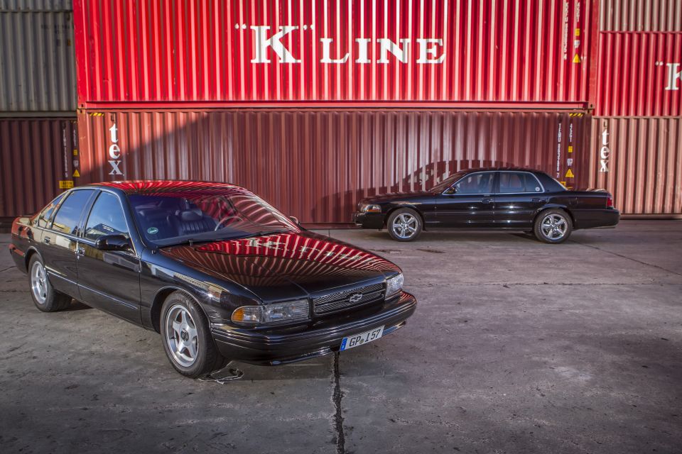 Two black sedans parked in front of red shipping containers. One is a Chevrolet Caprice, the other is a Ford Crown Victoria