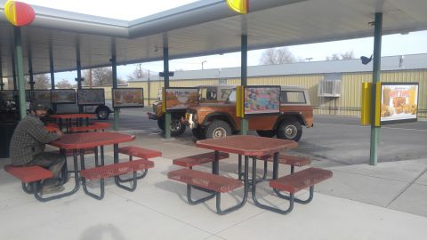 1971 Ford Bronco - An orange SUV is parked at a drive-thru with a man seated at a nearby picnic table.