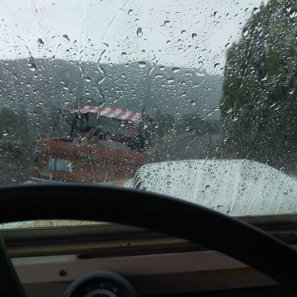 View through a rain-covered windshield at an orange construction vehicle with a red and white striped boom. Another vehicle is partially visible in the foreground