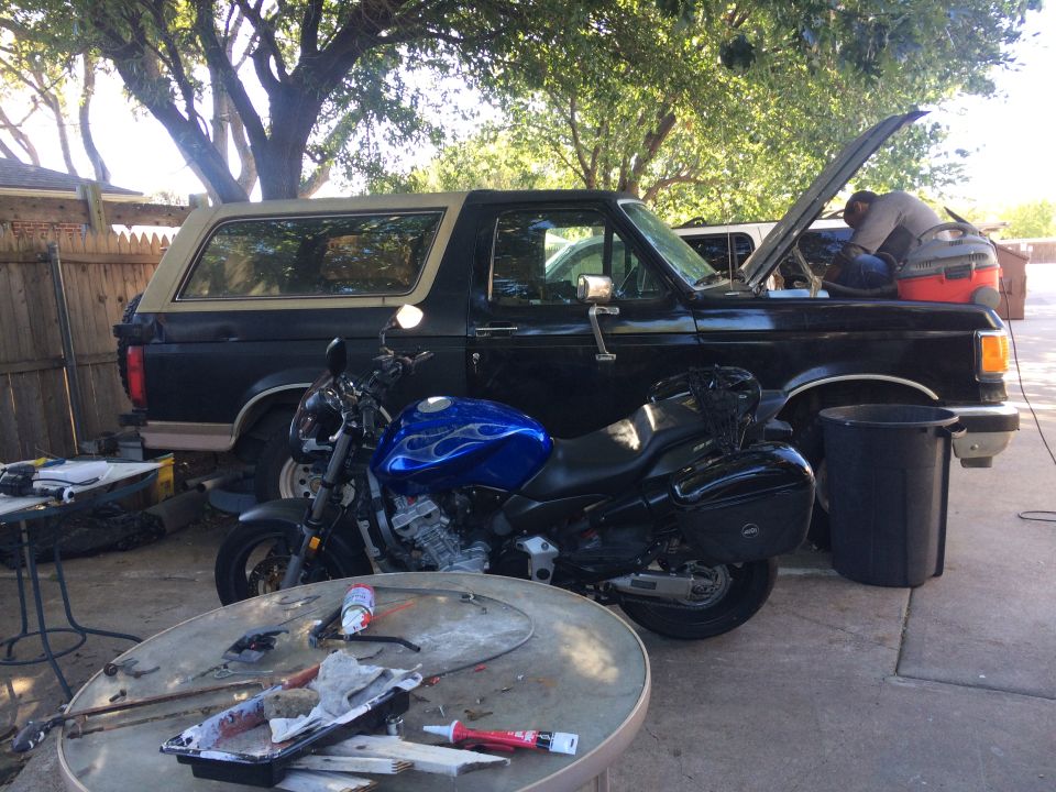 1989 Ford Bronco parked next to a blue motorcycle, with a person working on the Bronco's engine