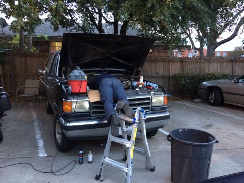1989 Ford Bronco - A person is working on the engine of a black truck with the hood open, using a ladder for access.