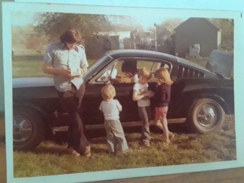 1994 Ford Bronco - A family gathers around a black 1967 Ford Mustang coupe, distinctive louvers on the rear quarter window.