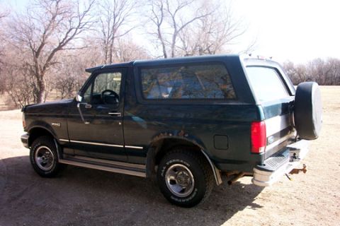1995 Ford Bronco - A dark green Ford Bronco is parked outdoors on a dirt lot with trees in the background.
