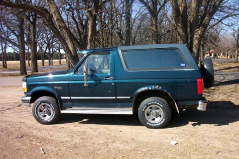 1995 Ford Bronco - Dark green two-door Ford Bronco full-size SUV photographed in profile from the driver's side, parked on a dirt lot among bare winter trees.…
