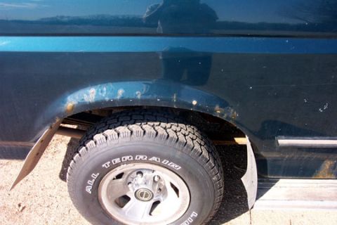 1995 Ford Bronco - Close-up of a blue truck's rear quarter panel and wheel opening, showing rust-through and bubbling paint along the arch lip with the mud fl…
