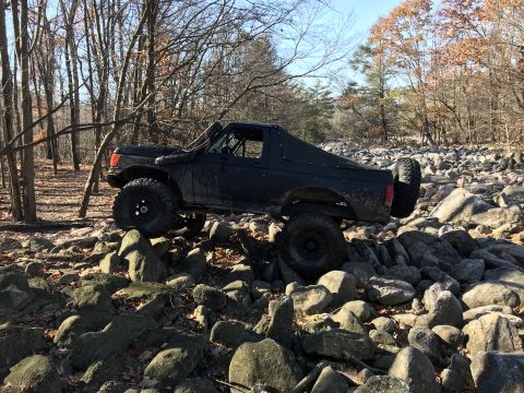 1989 Ford Bronco - Modified black SUV navigates a rocky terrain with a snorkel and spare tire visible.