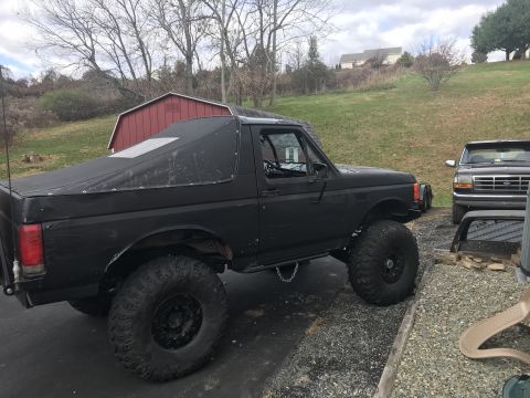 1989 Ford Bronco - This lifted 1987 Ford Bronco features a black soft top and is parked outdoors next to another Ford truck.