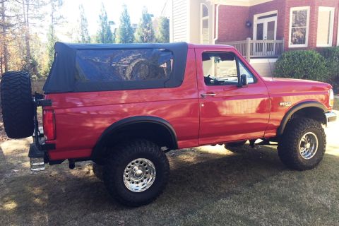 1994 Ford Bronco - Side view of a red 1994 Ford Bronco a soft top and a spare tire mounted on the rear.