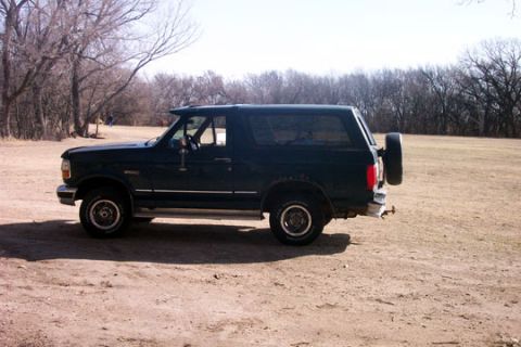 1995 Ford Bronco - 1995 Ford Bronco parked outdoors on a dirt surface with trees in the background.