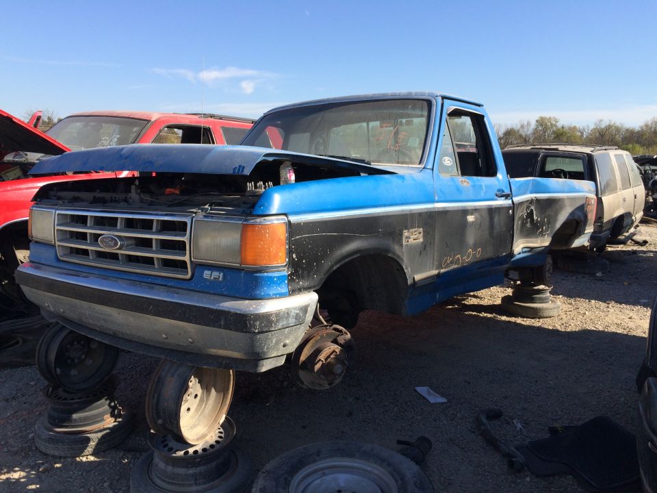 Blue and black Ford F-Series truck with hood up, missing wheels, at a junkyard. Another red vehicle and a tan SUV are in the background