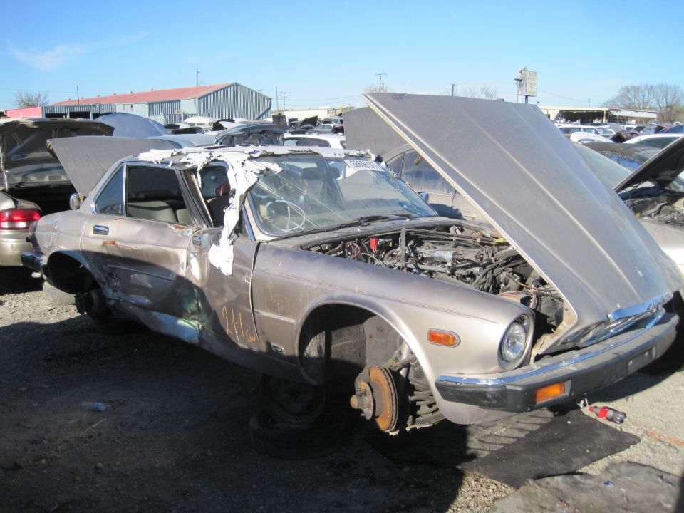 1985 Jaguar XJ6 with hood open, missing front wheels, and damaged roof in a junkyard