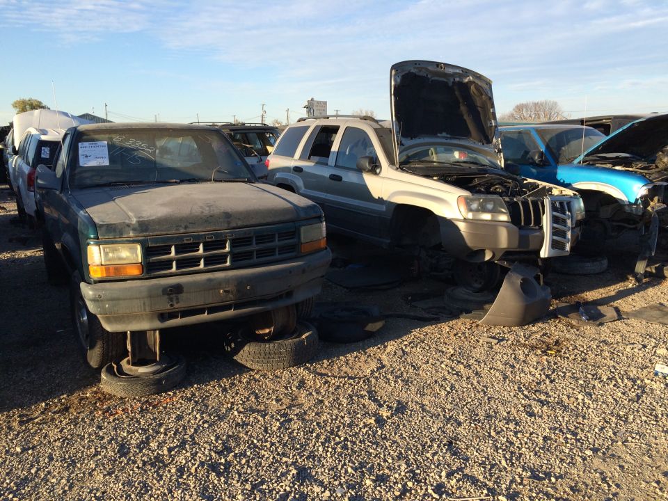 Junkyard scene with several vehicles, including a dark green Dodge pickup and a silver Jeep Grand Cherokee with its hood open