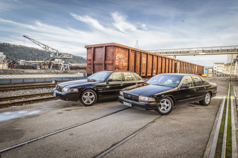 Two black sedans, one a 1996 Chevrolet Impala SS, parked side-by-side on train tracks near a freight train car