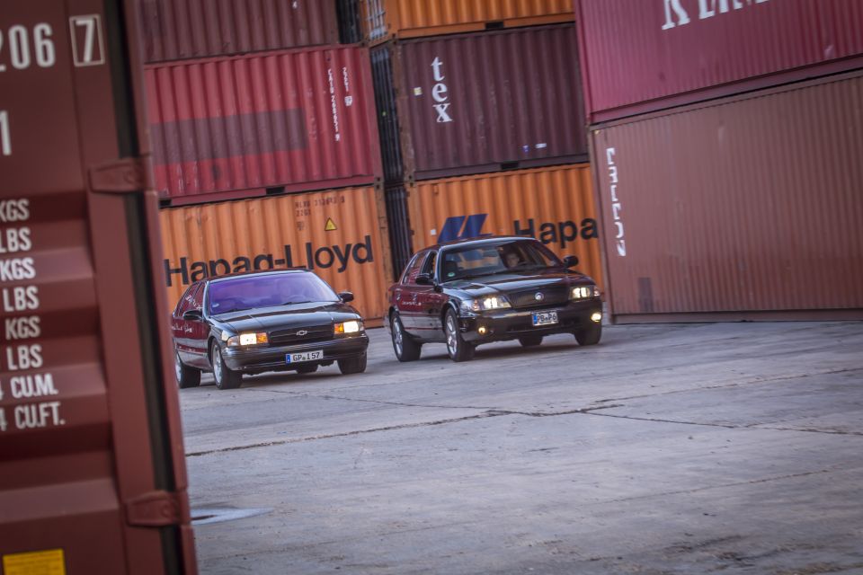 Two dark sedans, one with a license plate reading GRL157, drive past stacked shipping containers