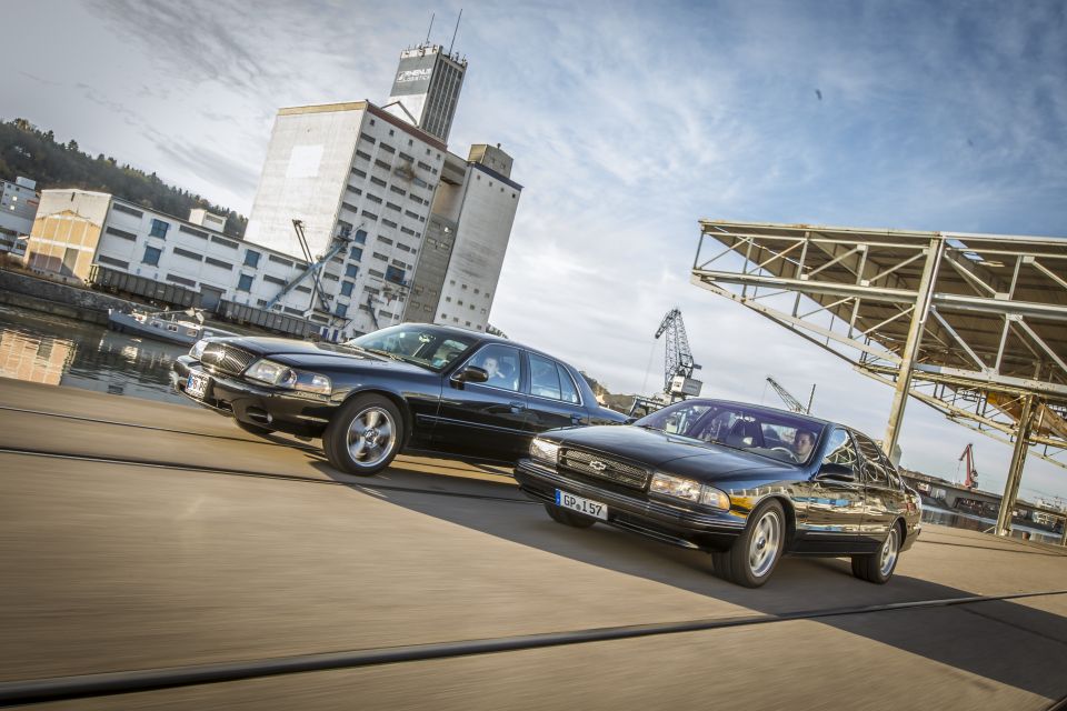 Two black sedans, one with a grille resembling a 1990s Chevrolet Caprice, drive on a paved surface near a waterfront industrial area